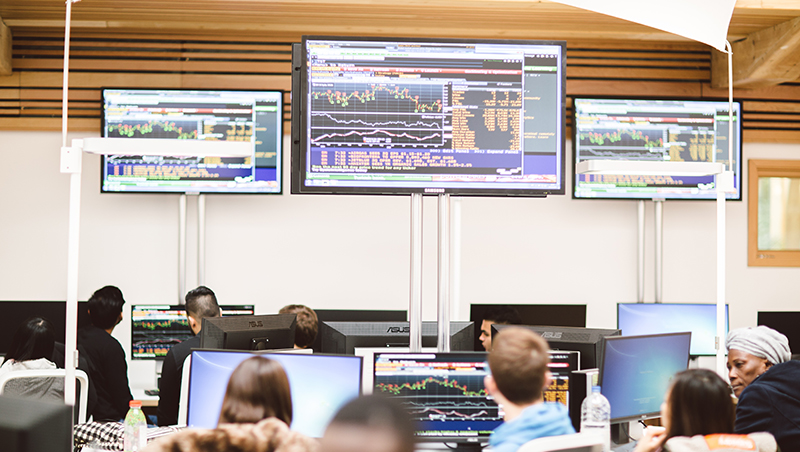 A wide angle shot of a class taking place in the Essex Business school trading floor with bloomberg data displaying on the large screens.
