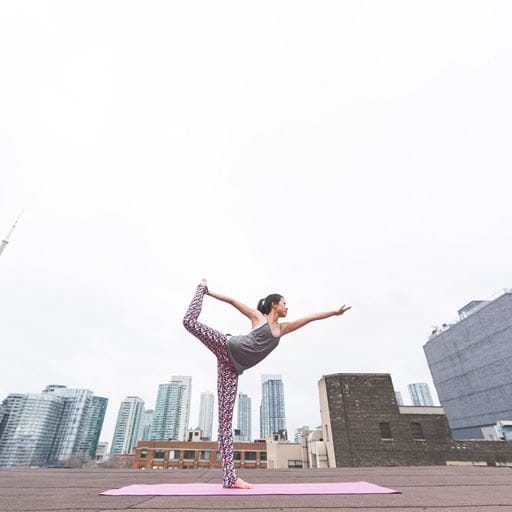 A woman standing outside on a pink mat, holding a yoga pose on one leg with her other foot behind her head. High-rise buildings are visible in the background.