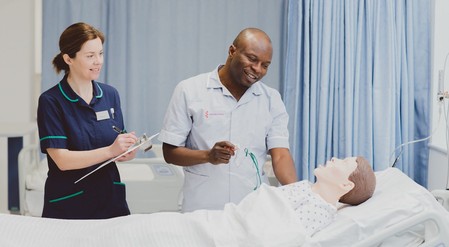 Kathy Burke is standing on the left holding a clipboard. To the right is a male nursing student holding an oxygen mask. In front of them is a Sim Man in a hospital bed in one of the School's mock wards.