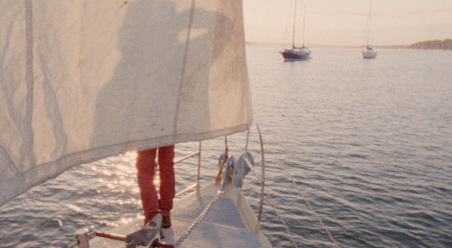 A person is seen in shadow behind a sail, standing on the deck of a boat, at sunset