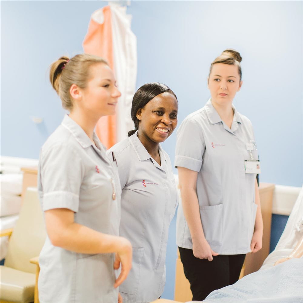 Three students in nurses uniforms standing next to a bed in a mock ward, listening to someone out of shot to the right of the photo.