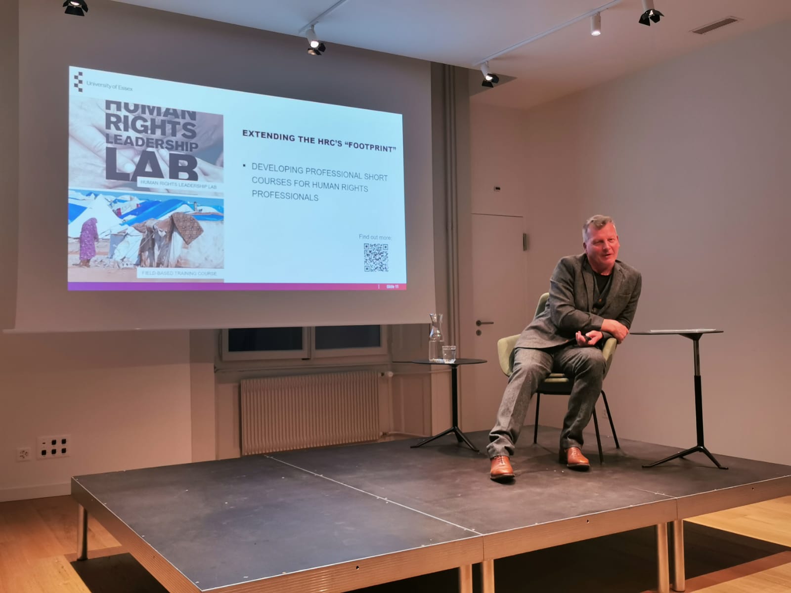 Dr Andrew Fagan, a white man in grey suit is sitting on a stage with a slideshow behind him with images of the posters for the short-courses offered by the HRC