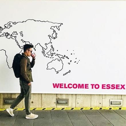 Student walking past a wall which has a world map and 'Welcome to Essex', student is drinking a coffee
