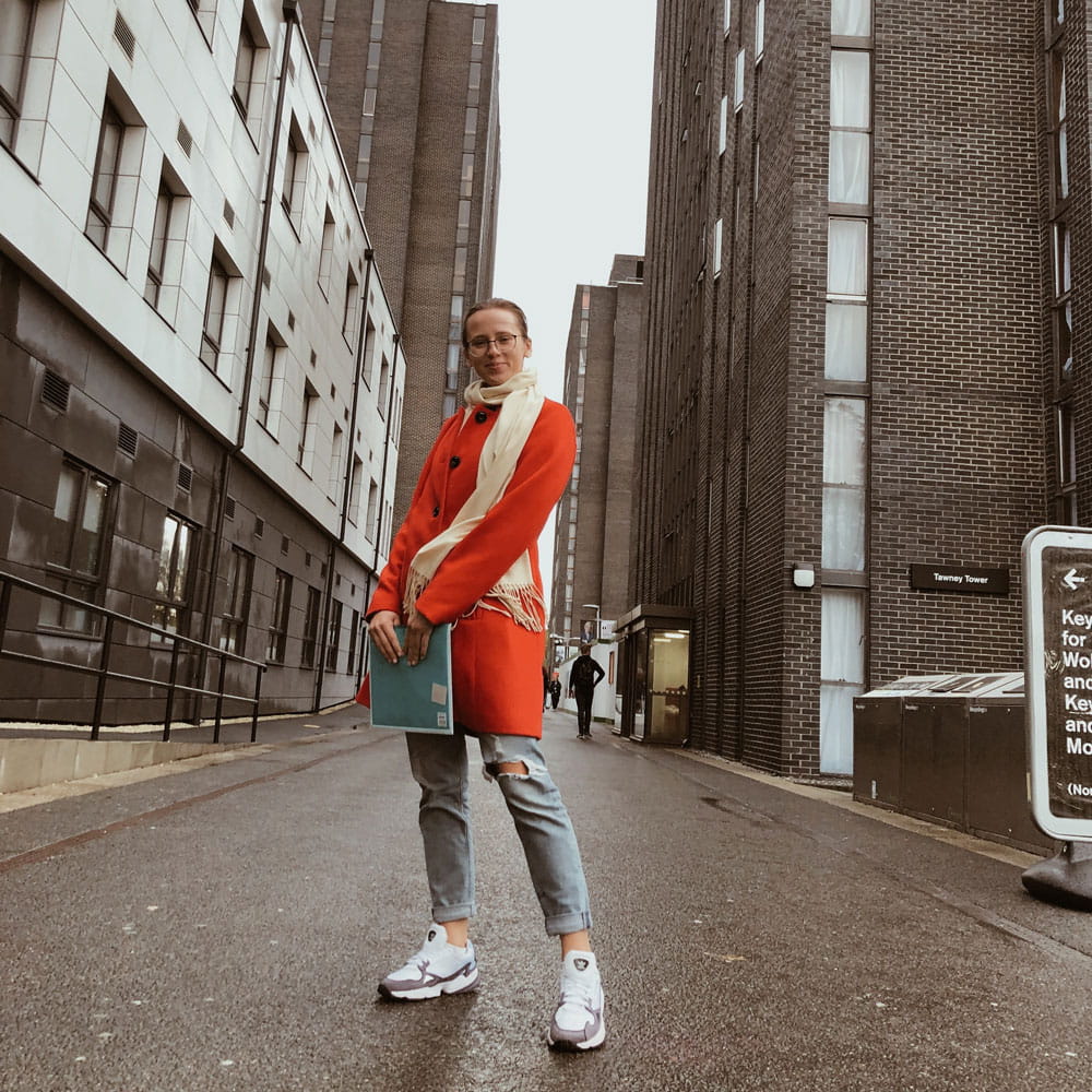 Student standing between The Towers in a red coat, holding a book