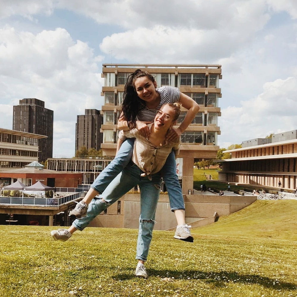Student on a piggy back with another student on grass infront of the library
