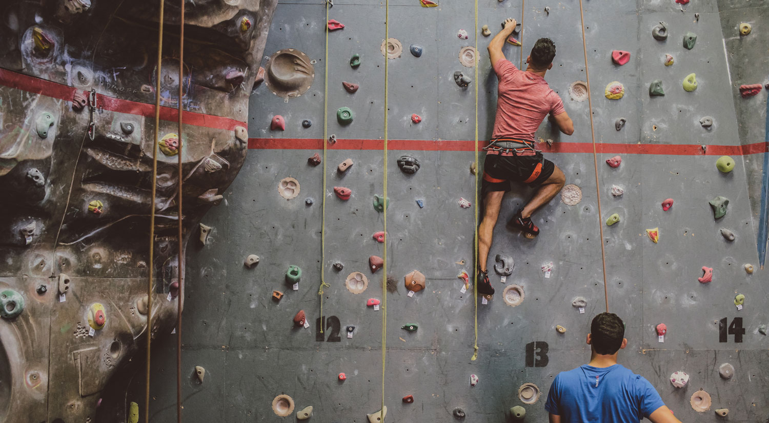 Two people climbing a climbing wall, one on the wall, the other guiding from below.