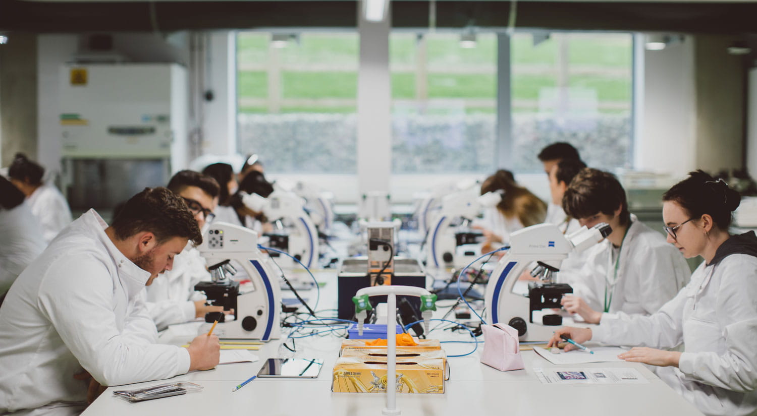 Students sitting in a lab wearing white coats looking into microscopes)