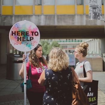 Student ambassador holding a sign that says 'here to help' whilst talking to two visitors on Square 4