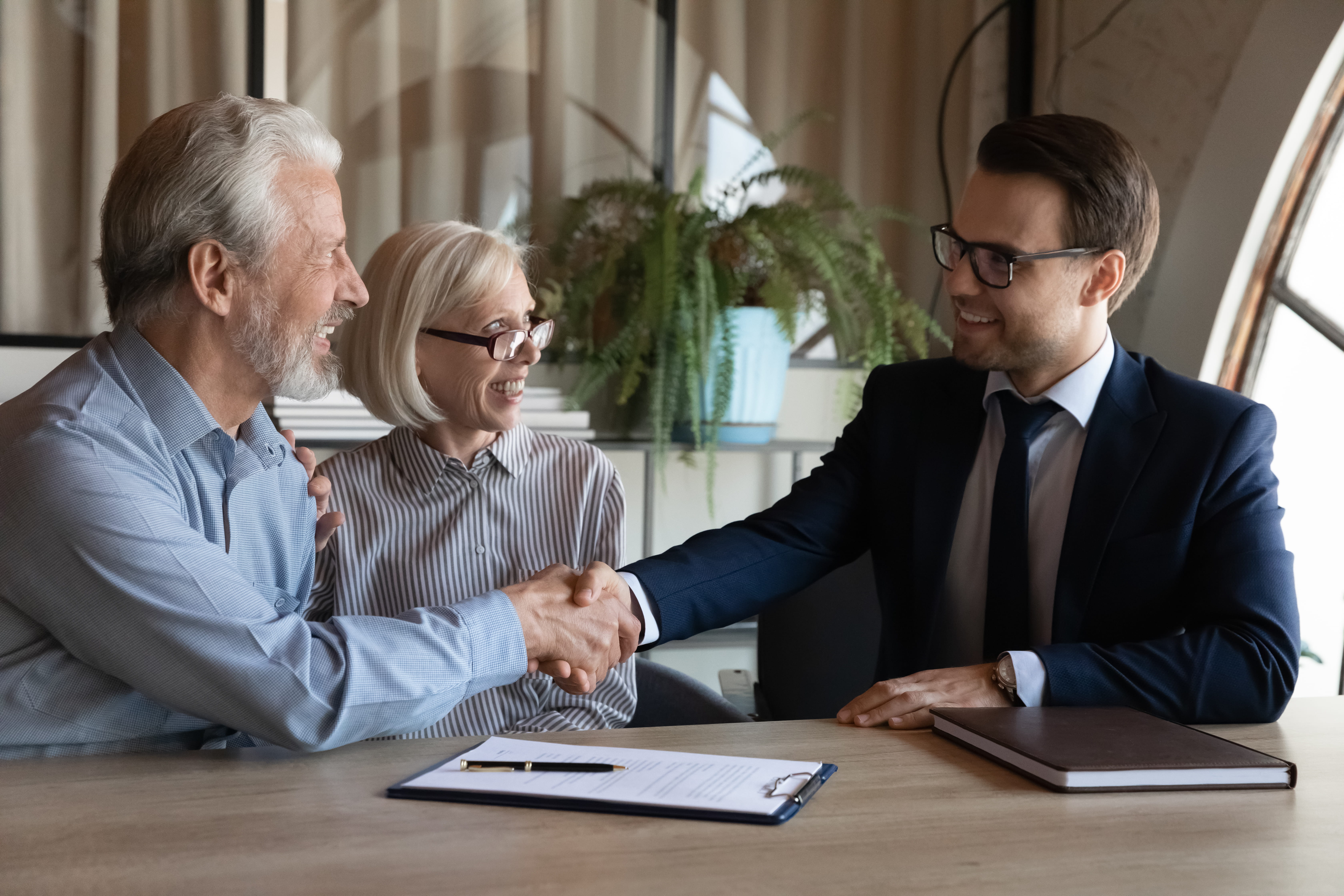 A couple of A man and a woman shake hands with a solicitor wearing a black suite and glasses. They are sitting on a table and a contract a pen and a book is on top of the table.