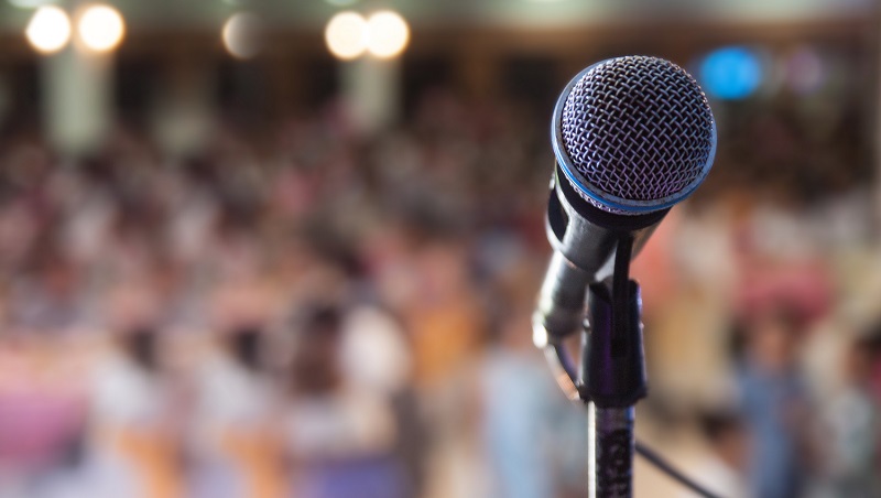A close up of a microphone on a stand with a packed conference hall in the background.