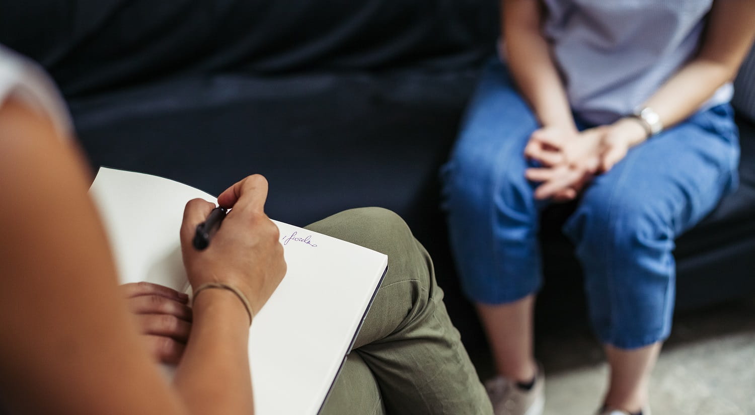 A  therapist takes notes in a book from a patient sitting in opposite chair.