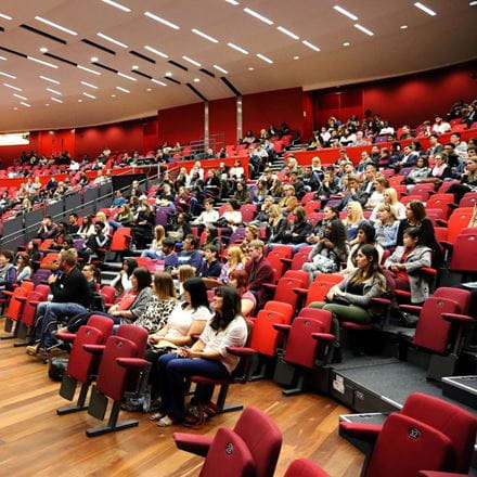 Lots of people sitting in a busy lecture hall with red seats