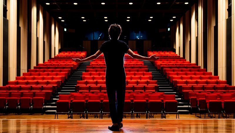 A young actor on a stage with hands held out facing an empty auditorium.)