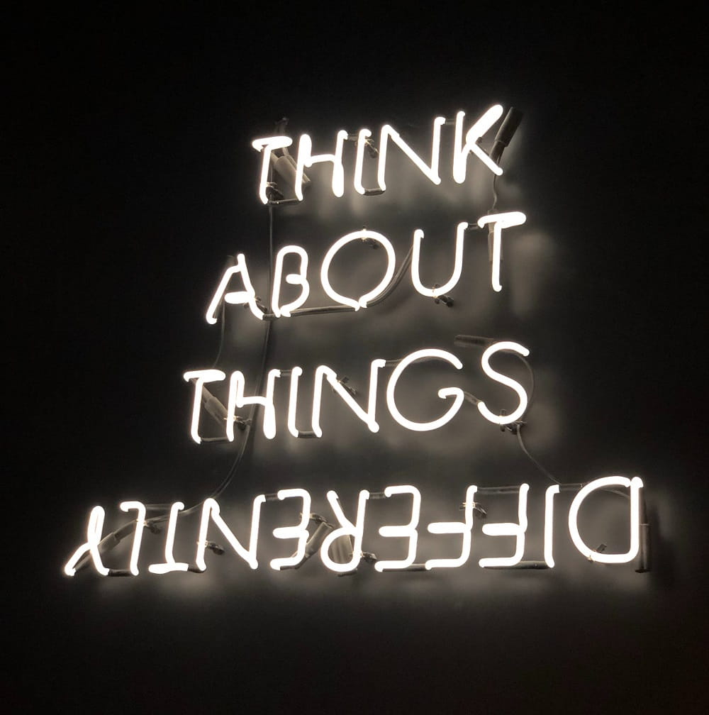 Black background with white neon stacked sign which says 'think about things differently' 