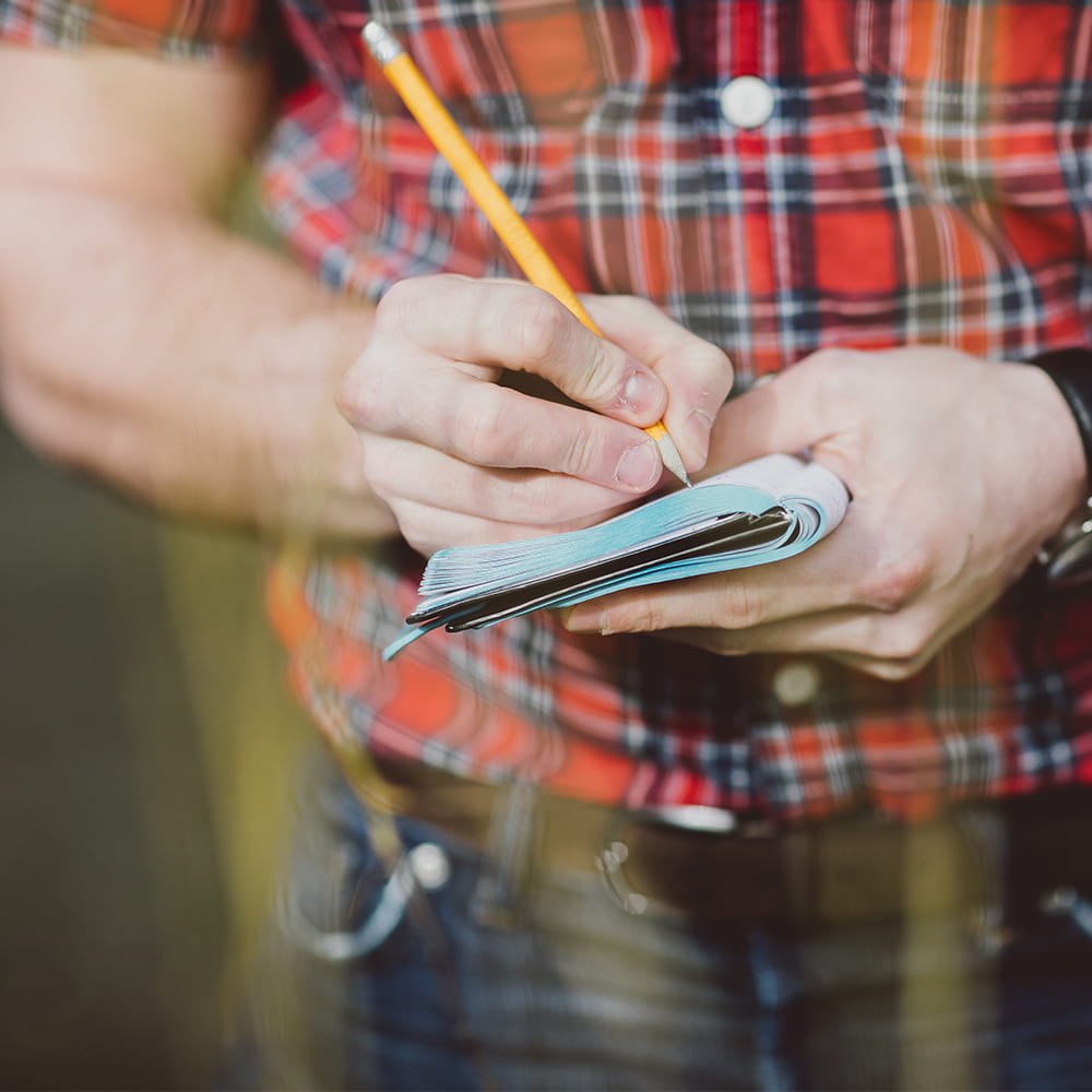 Man wearing a red plaid shirt and writing in a notebook outside.