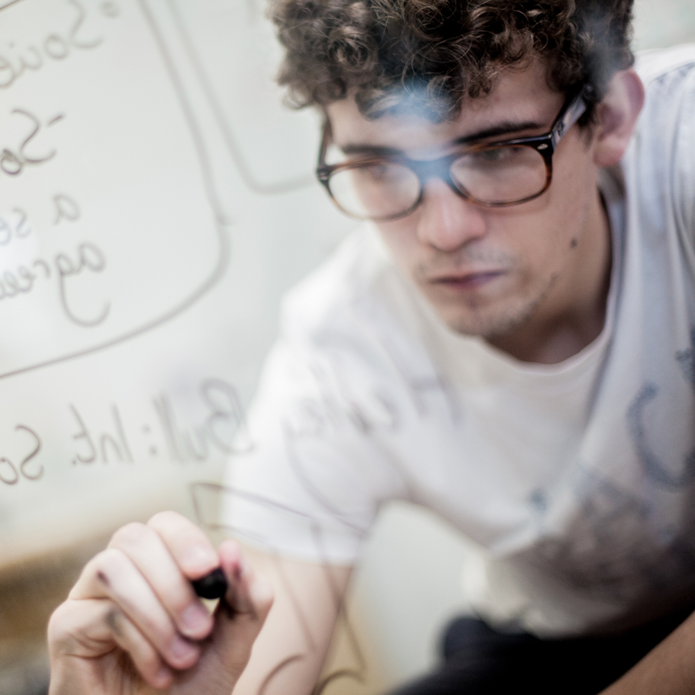 A student wearing glasses, writing equations in marker pen on a glass surface.