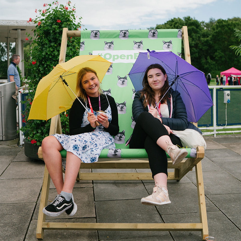 Two girls sitting on a giant armchair at an Open Day, each holding an umbrella