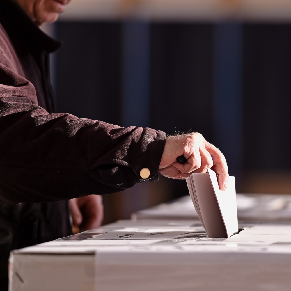 A man in a dark winter coat holding a ballot paper as he drops  it in the ballot box
