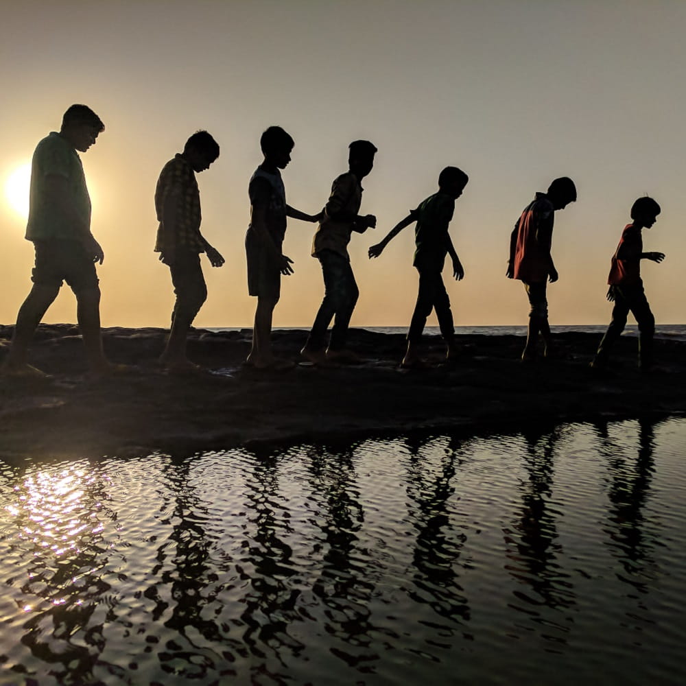 A line of young children walking on a beach with the sun in the background.