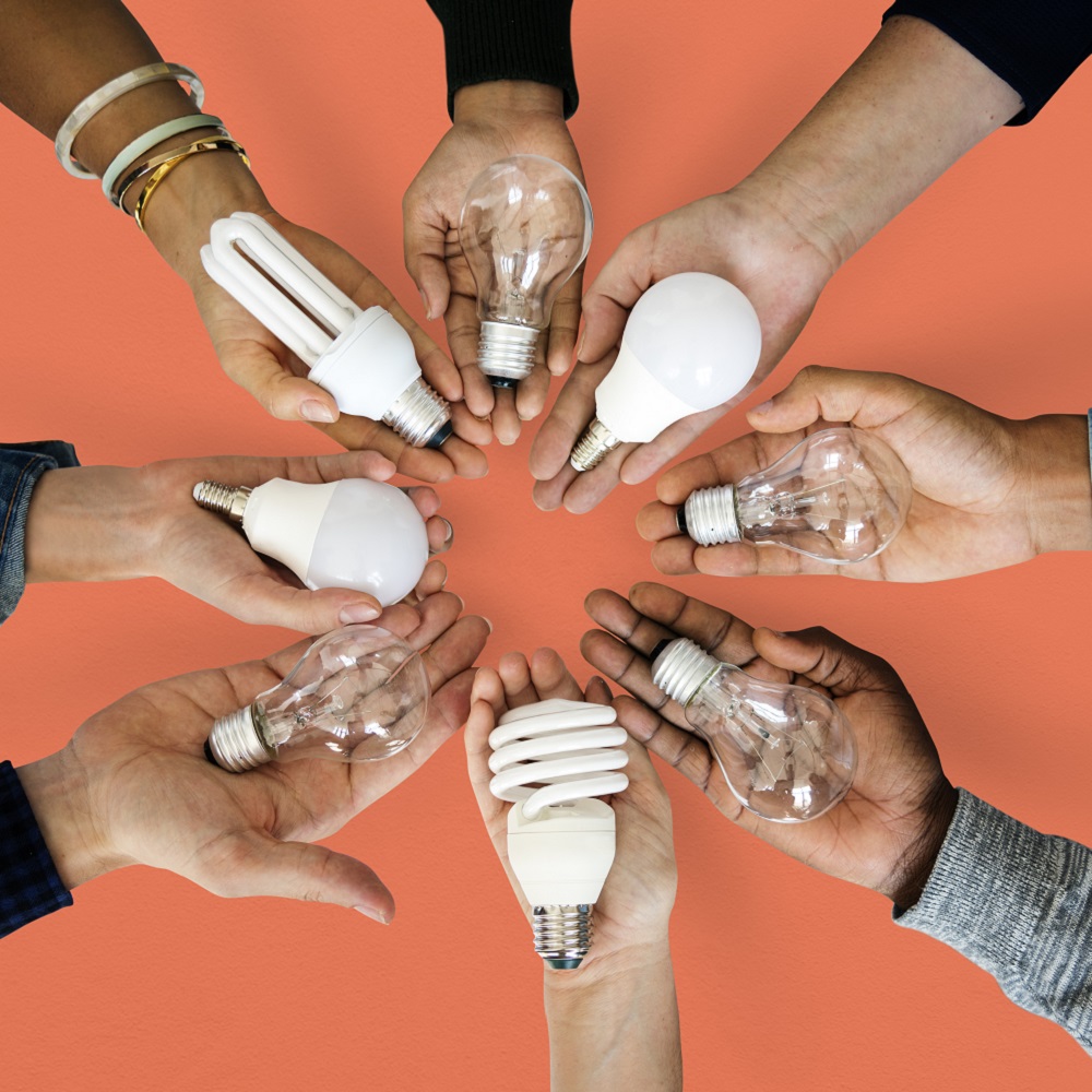 Eight people with their hands held out with different styles of light bulb in the palms of their hands on an orange background.