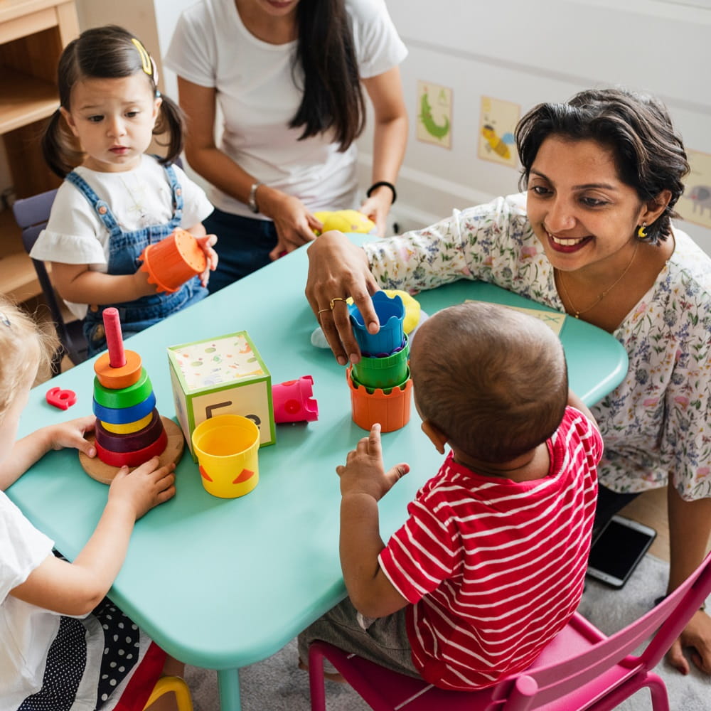 A group of young toddlers sat at a table with a teacher, playing with pre-school toys.