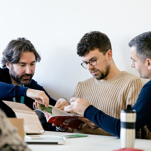 Three male MBA students sit at a desk in discussion together.