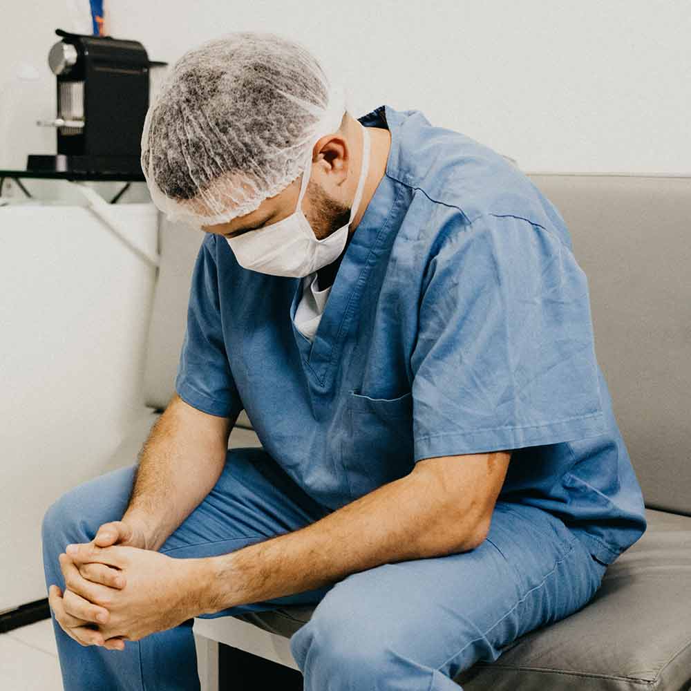 Dejected doctor, wearing blue scrubs and face mask, hanging his head