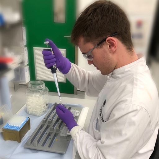 Student Joe Wilmshurst working in a lab, wearing a white coat and purple gloves