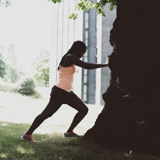 Black woman wearing a pale pink top and black shorts doing leg stretch exercises against a tree in a green space