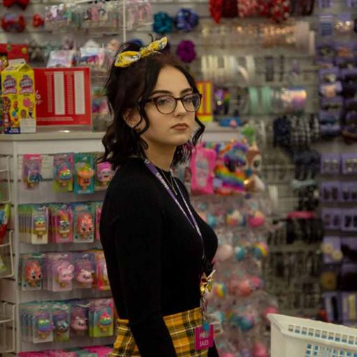 Young woman wearing a black top, yellow tartan skirt and black-rimmed glasses standing in accessory and jewellery shop Claire's