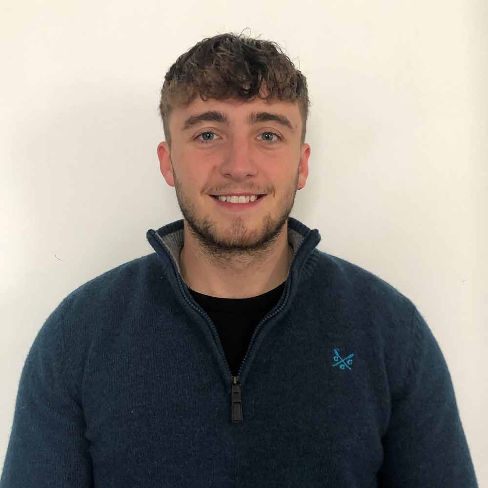 Joseph Barker, wearing a dark blue collared jumper and standing against a white wall