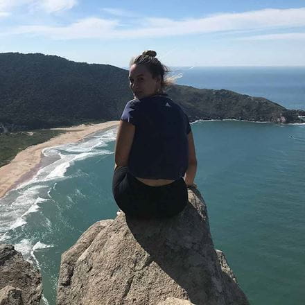 Nina Beckmann Gámez sitting on a high peak above a beach and blue sea, looking back towards the camera