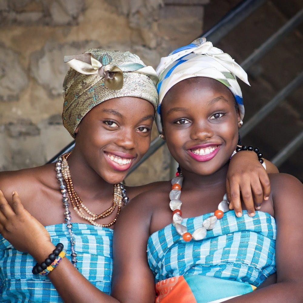 Two smiling African women wearing headscarves, blue dresses and brightly coloured jewellery.