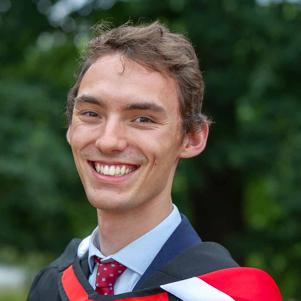 Lewis Smith, smiling in front of a woodland backdrop, wearing a suit and tie with his Essex graduation robes just visible