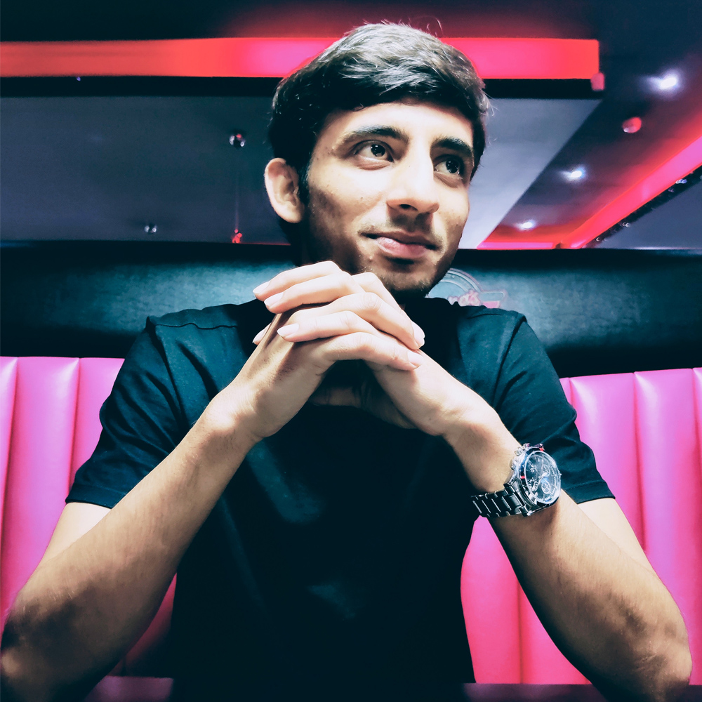 A young gentleman sits against a pink and black background. He smiles whilst looking away from camera.