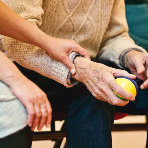 A close up of an elderly man's hands. He is holding a ball in one hand and the hand of a younger care worker is resting on his wrist
