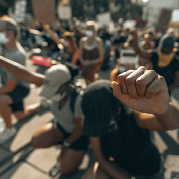 A group of protesters, mostly out of focus. In the foreground two young Black people wearing baseball caps kneel, each with a fist raised. The fist on the bottom right is in focus.
