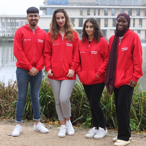 4 individuals standing infront of the lake near the Silberrad Student Centre. They are wearing Residence Life iconic red hoodies and smiling at the camera. 