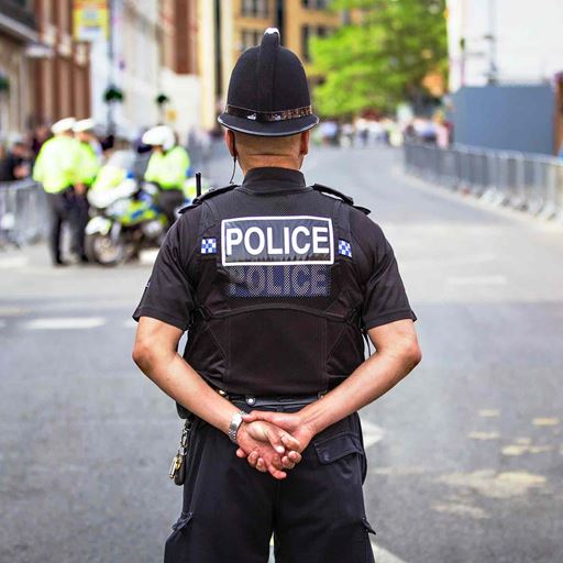 A male police officer, seen from behind, stands in the middle of a road with his hands clasped behind his back and the word 'Police' clearly visible on the back of his uniform