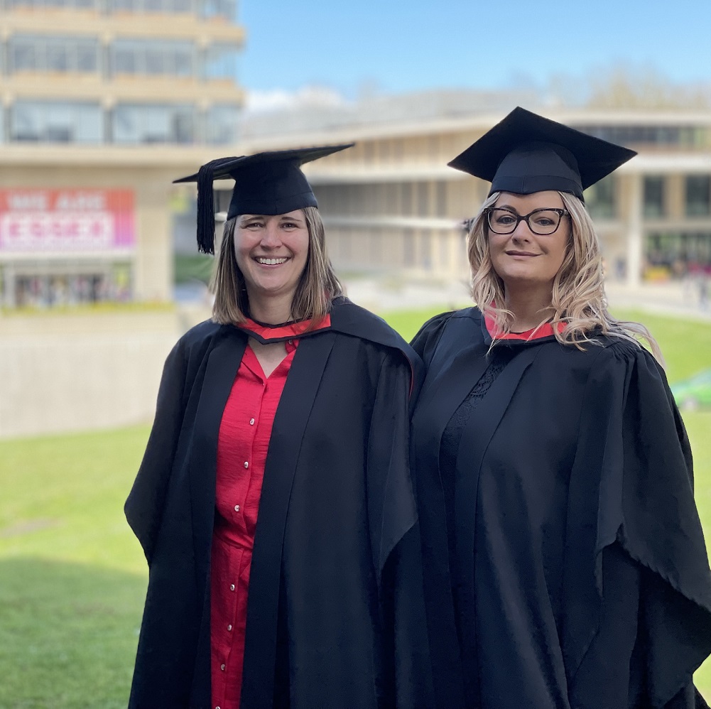 photo of two female graduates in robes outside on grass in front of  Essex University Library