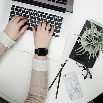 A pair of hands over a laptop keyboard, with a small plant, a pair of glasses and a phone on the right hand side.
