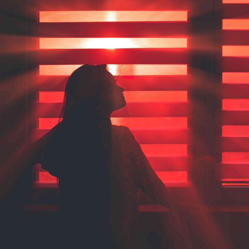A silhouetted young woman is seen standing in front of a window with blinds in a hazy, red light