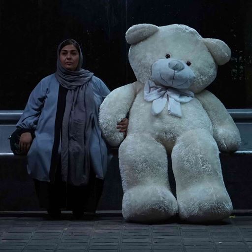 A depressed looking woman, of middle eastern origin, sits at a bus stop with a human-size cream teddy bear sitting next to her.  Still from Tehran: City of Love courtesy of Here and There Productions, 2018