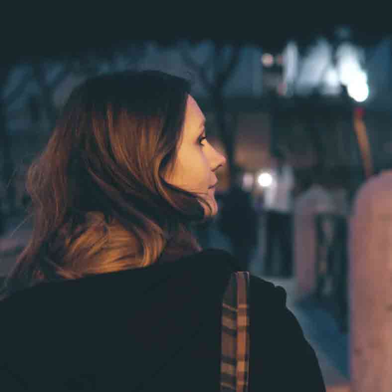 A young woman, walking at night, turns her head slightly as if listening for something behind her