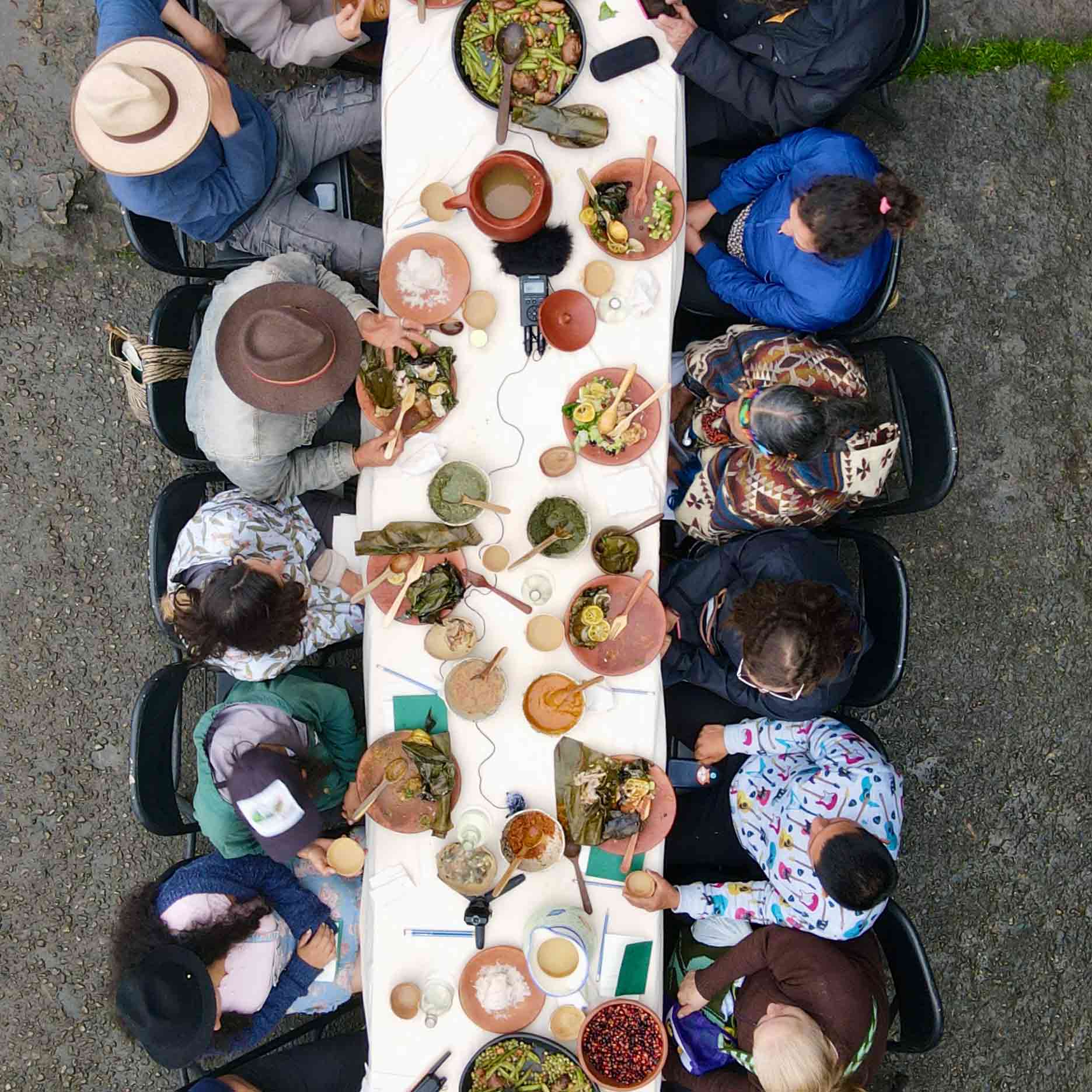 A long dining table around which a group are seated, seen from above and laden with food