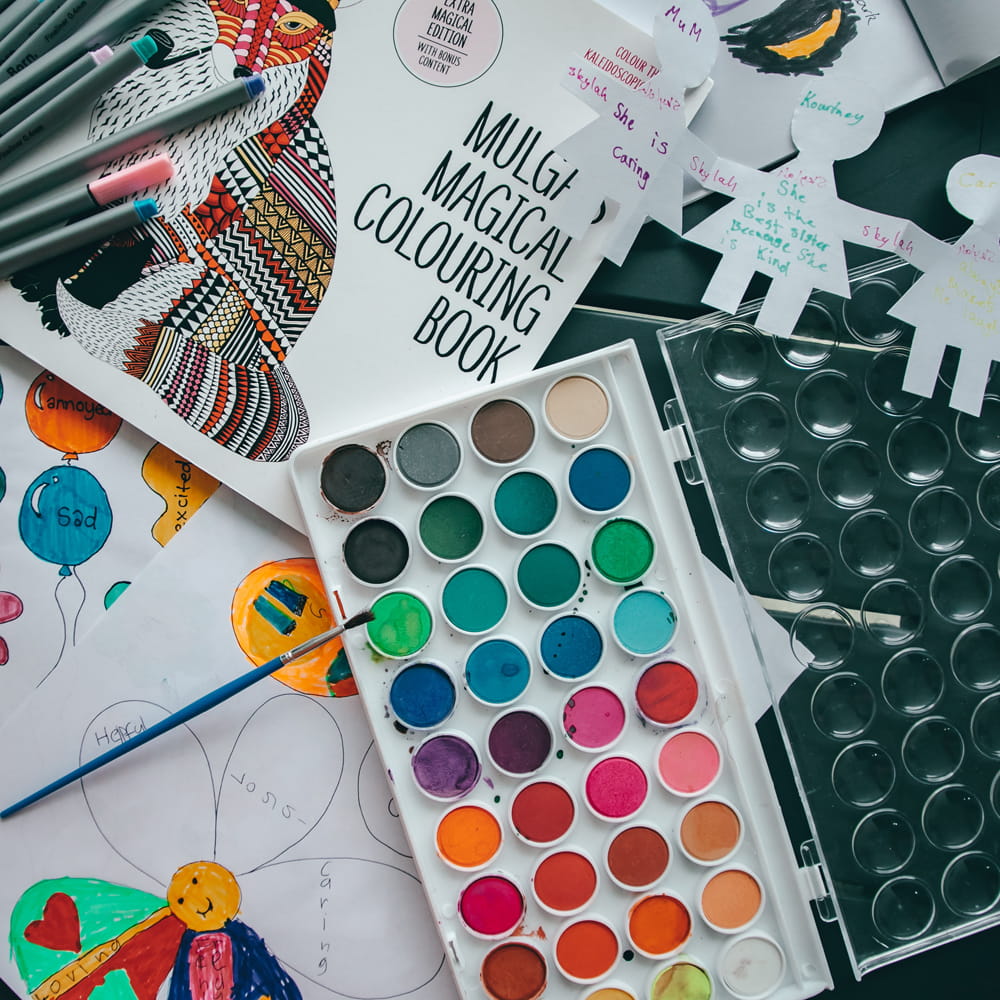 A tray of paints are on a table, with paintbrushes, pens and colouring sheets all scattered around nearby. 