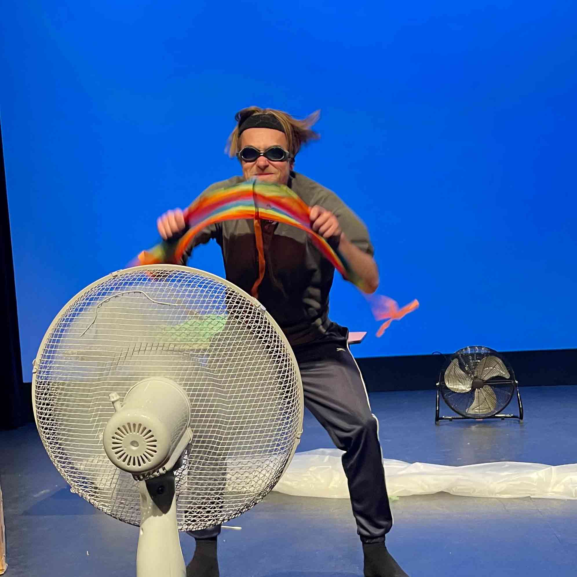 A young male stands, wearing goggles, in front of a large fan