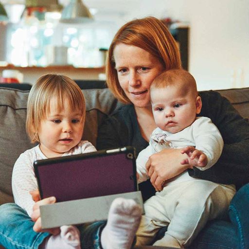 Woman sitting with a baby on her lap and a toddler next to her looking at an iPad