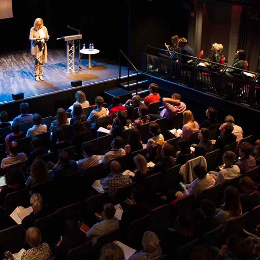 View across an audience, sitting in an auditorium as a woman on stage delivers a presentation