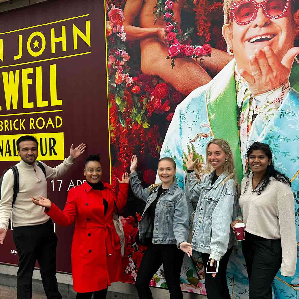 Five students, looking happy, stand in front of a giant Elton John poster board